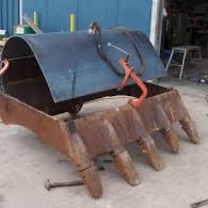 A rusty excavator bucket with sharp teeth, sitting on the ground with a blue industrial building in the background.